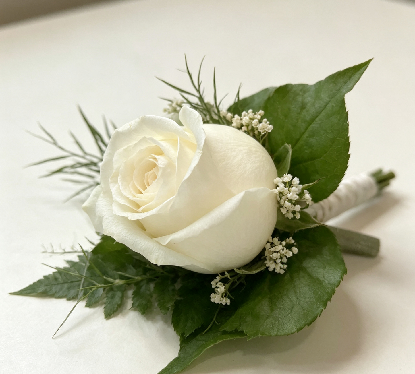 A white rose boutonniere floral arrangement with green leaves and white rose. White wrist corsage and buttonhole bundle for weddings and formal events