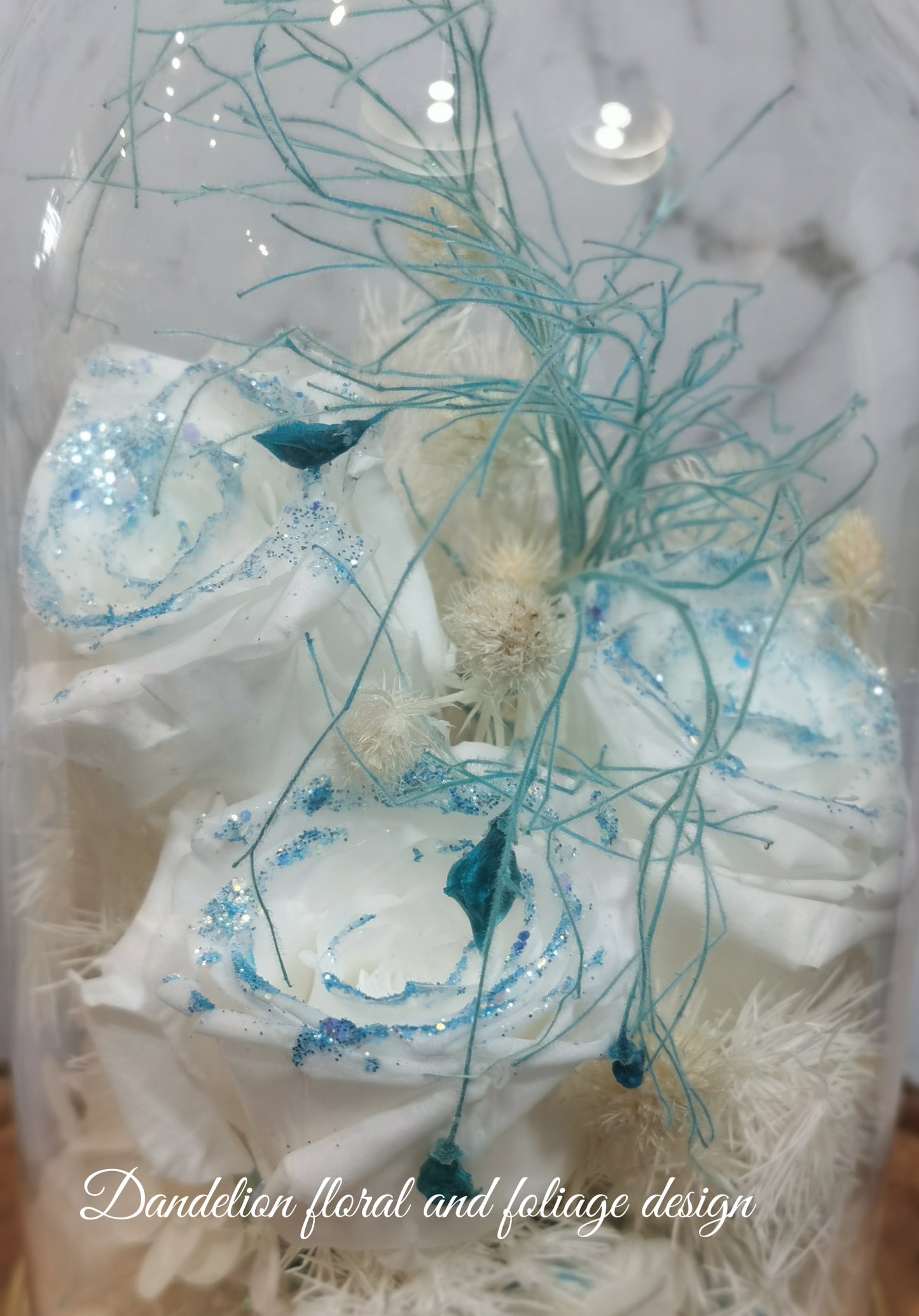 A handmade dome-shaped arrangement of preserved flowers in shades of blue, white, and beige, with a sparkling blue ribbon-like stem, set on a wooden stand.