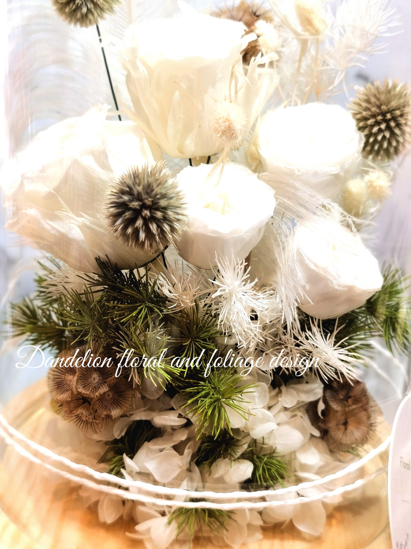 A preserved flower dome cloche arrangement with creamy feather-like flowers in a clear dome on a wooden stand.