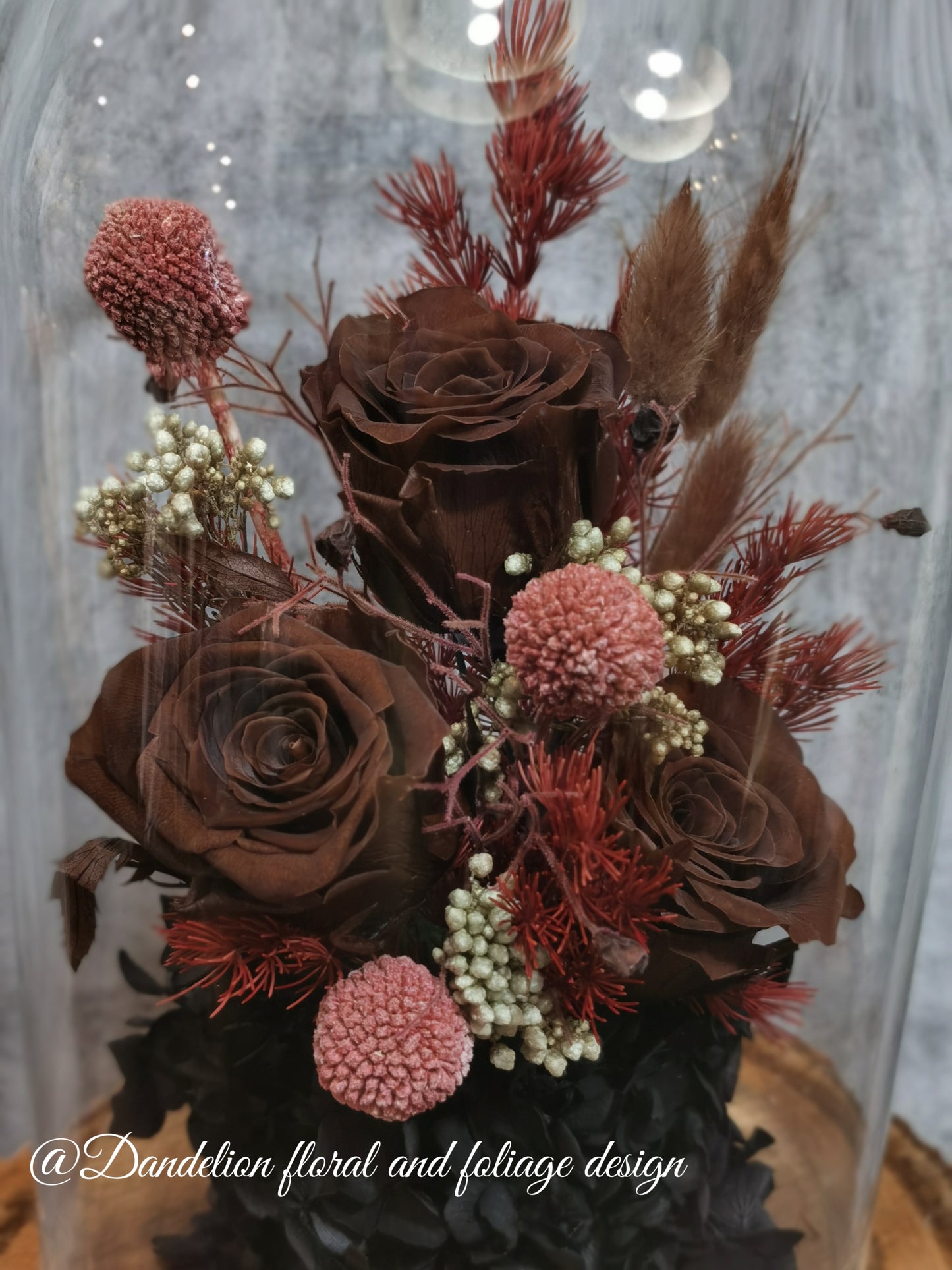 A handmade everlasting preserved rose and red flower dome cloche arrangement under a floral archway, displayed on a wooden stand.
