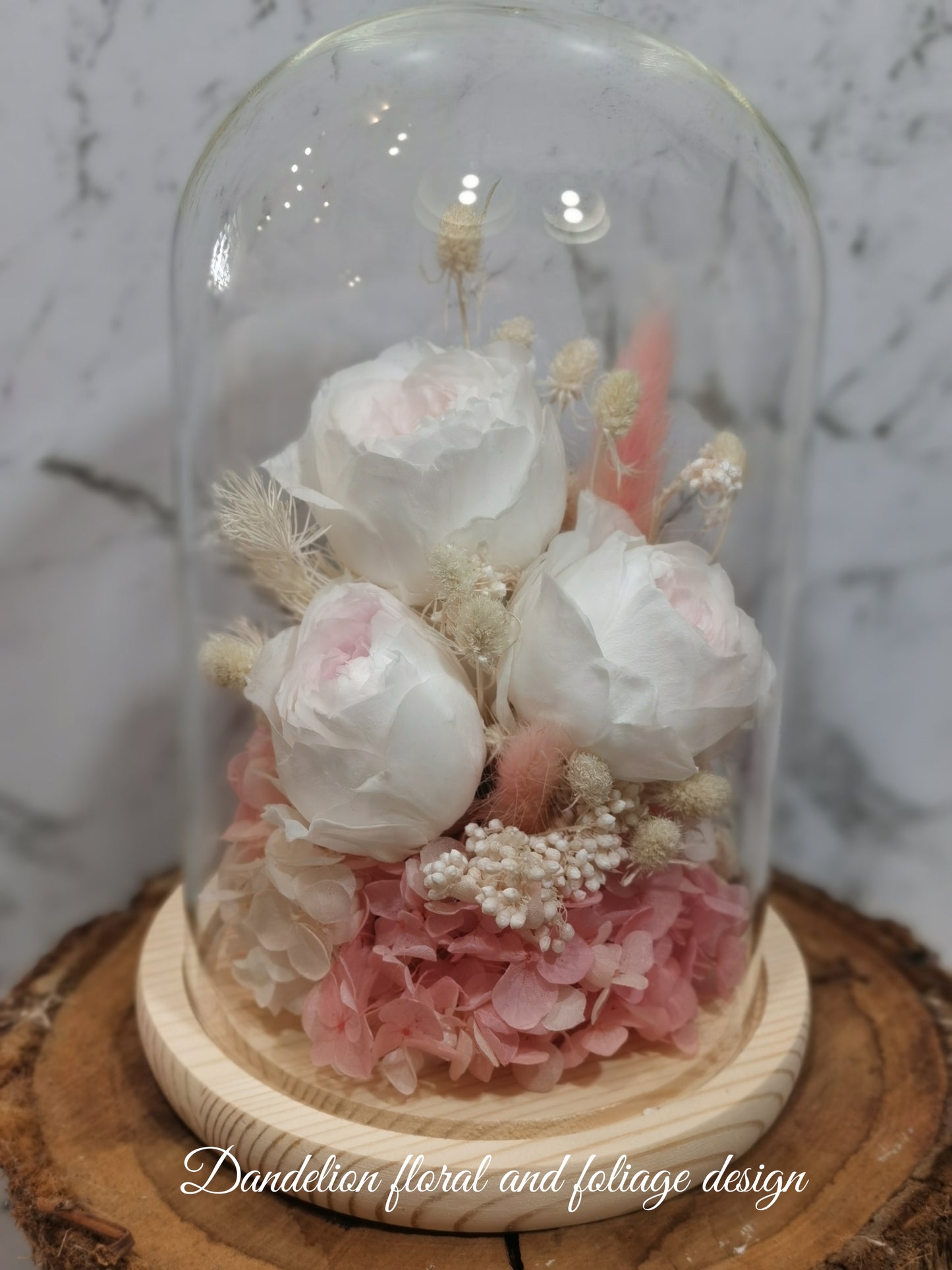 A handmade everlasting preserved white rose and pink flower dome cloche arrangement under a floral archway, displayed on a wooden stand.