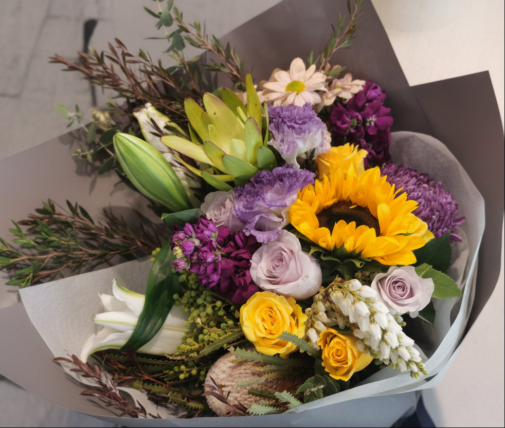 A bouquet of flowers with a mix of seasonal blooms and foliage, arranged and held by a mannequin. Bright colour bouquet using seasonal blooms complimented with seasonal foliage. Vibrant bouquet with sunflower, roses, lilies, natives and seasonal blooms in bright yellow and purple tones.