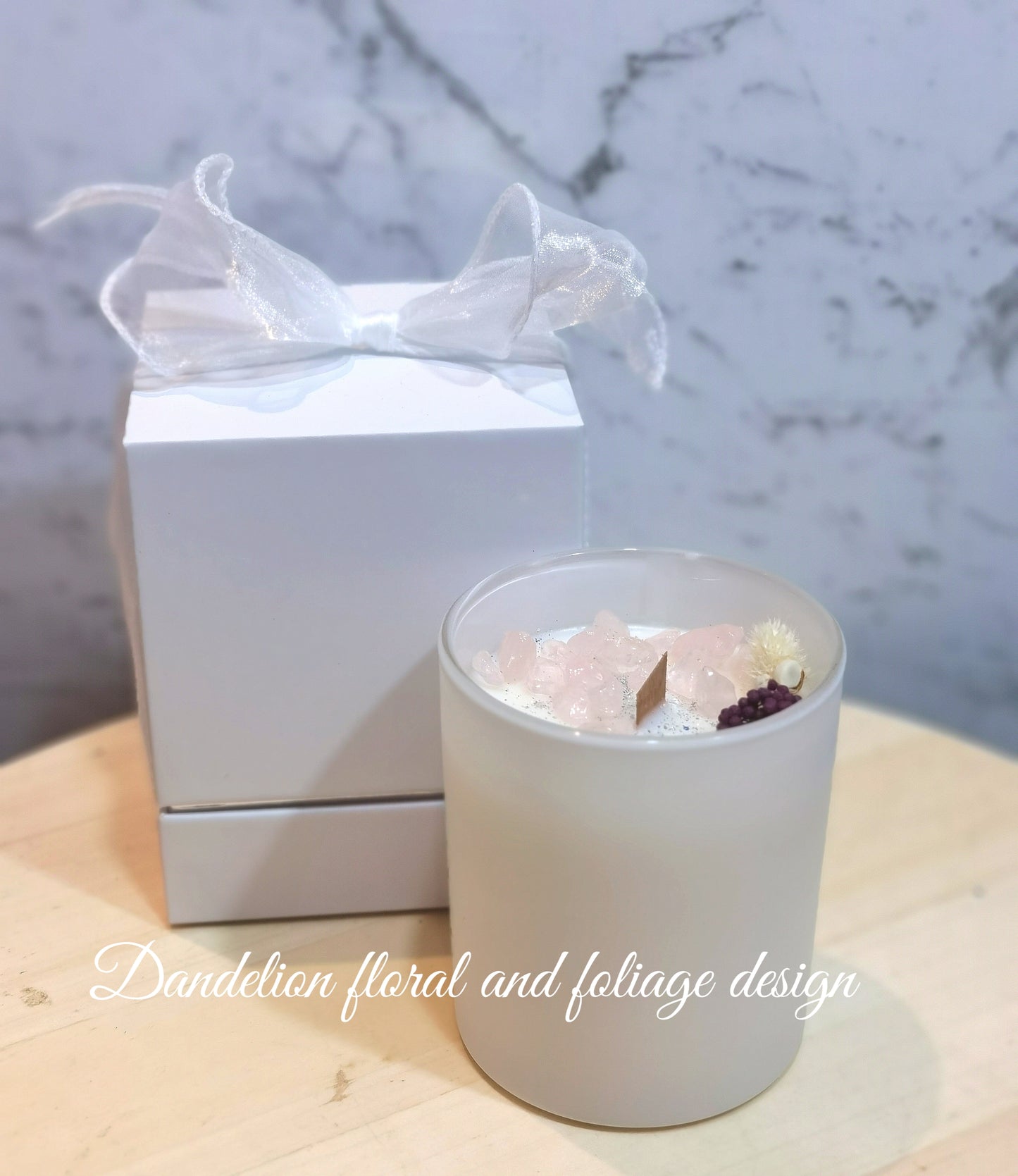 A contemporary white glass jar soy candle with a white lid, decorated with a white bow and placed next to a whitebox with a pink flower design.