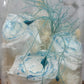 A handmade dome-shaped arrangement of preserved flowers in shades of blue, white, and beige, with a sparkling blue ribbon-like stem, set on a wooden stand.