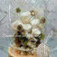 A preserved flower dome cloche arrangement with creamy feather-like flowers in a clear dome on a wooden stand.