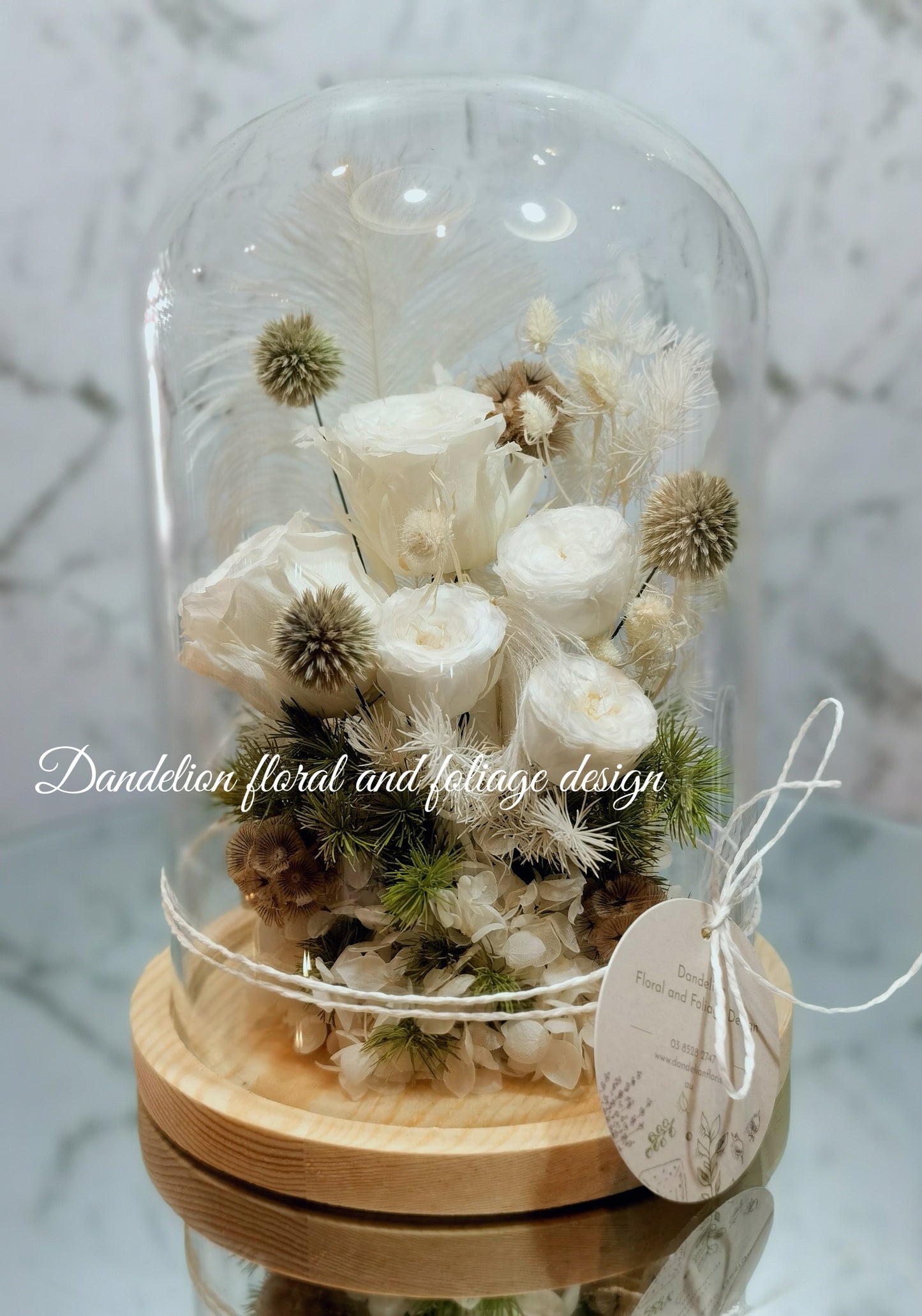 A preserved flower dome cloche arrangement with creamy feather-like flowers in a clear dome on a wooden stand.