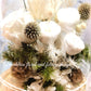 A preserved flower dome cloche arrangement with creamy feather-like flowers in a clear dome on a wooden stand.