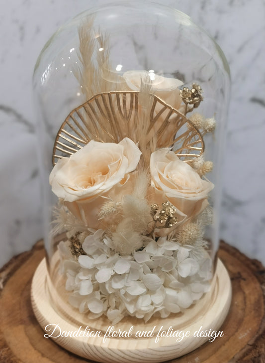 A preserved flower dome cloche arrangement with gentle apricot-colored flowers, bead embellishments, and a gold-tone frame, displayed on a wooden stand.