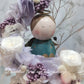A handmade everlasting preserved flower dome cloche arrangement with a mini angel doll seated under a floral archway, displayed on a wooden stand.