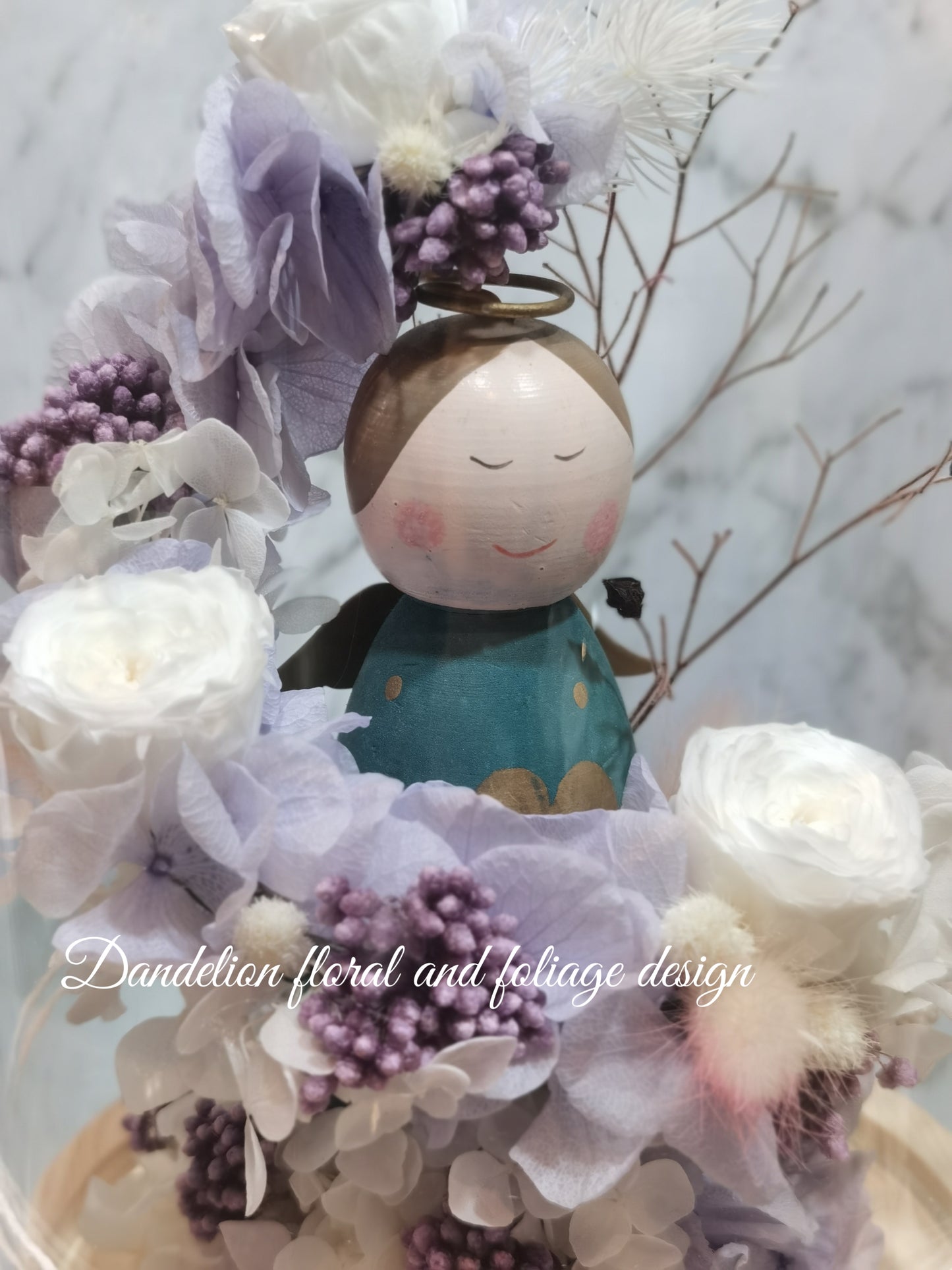 A handmade everlasting preserved flower dome cloche arrangement with a mini angel doll seated under a floral archway, displayed on a wooden stand.
