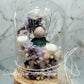 A handmade everlasting preserved flower dome cloche arrangement with a mini angel doll seated under a floral archway, displayed on a wooden stand.