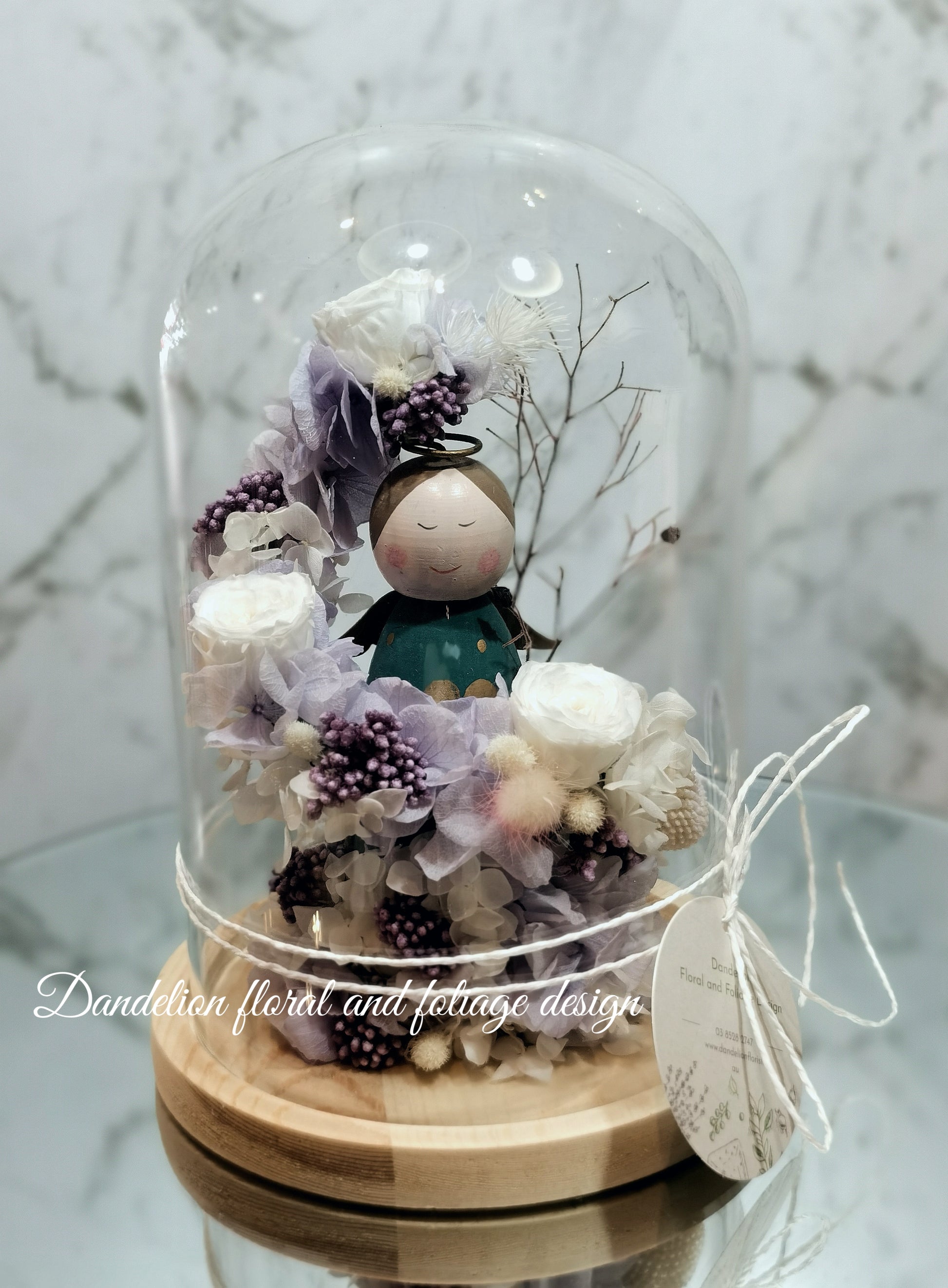 A handmade everlasting preserved flower dome cloche arrangement with a mini angel doll seated under a floral archway, displayed on a wooden stand.