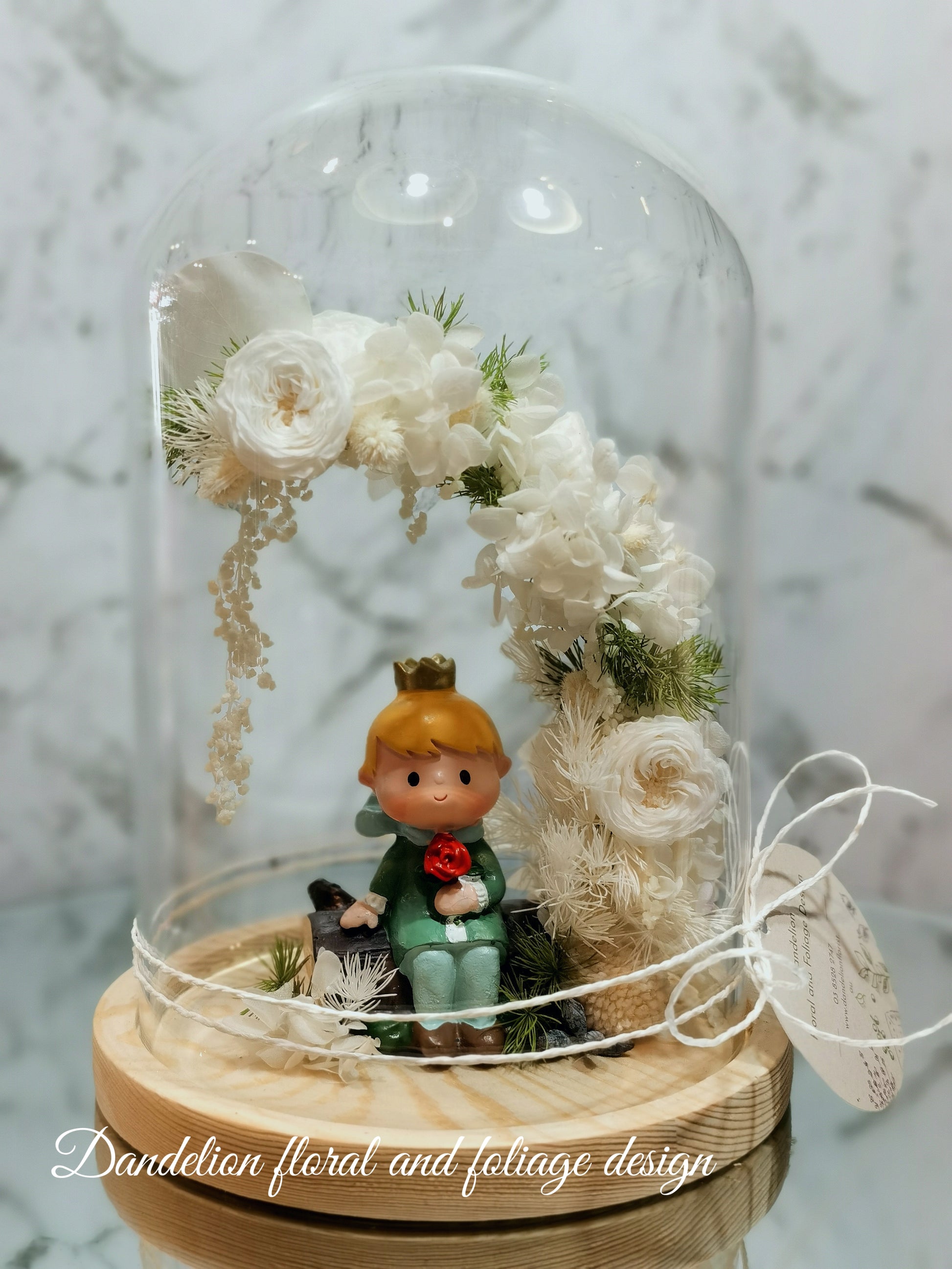 A handmade everlasting preserved flower dome cloche arrangement with a mini doll seated under a floral archway, displayed on a wooden stand.