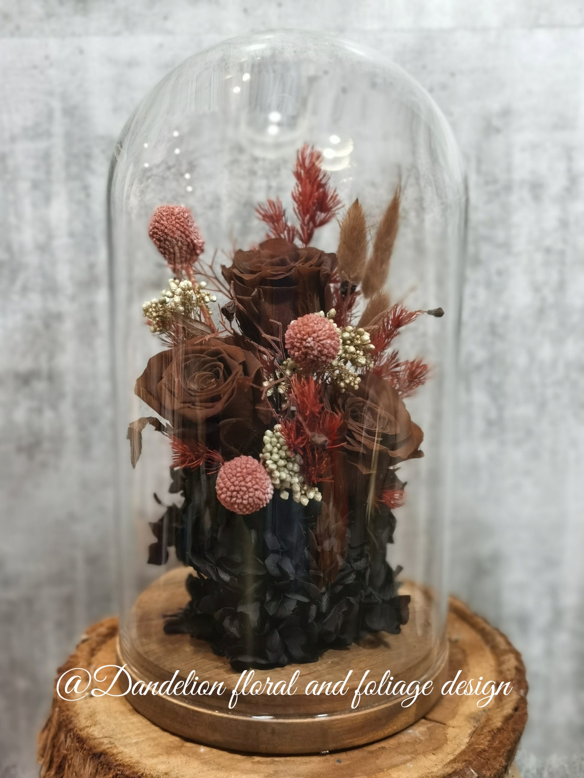 A handmade everlasting preserved rose and red flower dome cloche arrangement under a floral archway, displayed on a wooden stand.