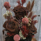 A handmade everlasting preserved rose and red flower dome cloche arrangement under a floral archway, displayed on a wooden stand.