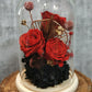 A handmade everlasting preserved red rose and pink flower dome cloche arrangement under a floral archway, displayed on a wooden stand.