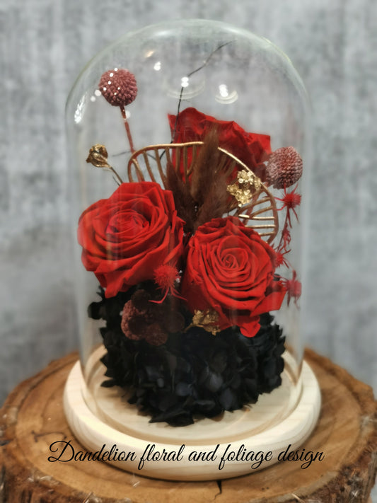 A handmade everlasting preserved red rose and pink flower dome cloche arrangement under a floral archway, displayed on a wooden stand.