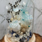 A handmade dome-shaped arrangement of preserved flowers in shades of blue, white, and beige, with a sparkling blue ribbon-like stem, set on a wooden stand.