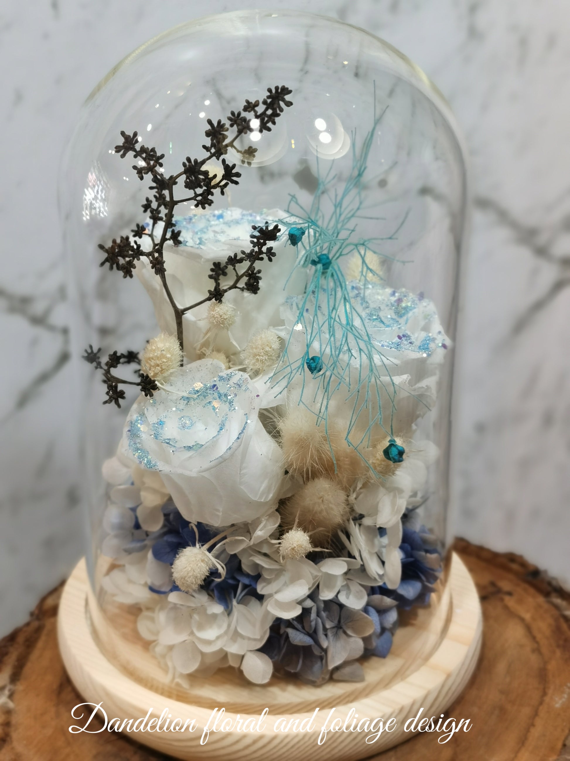 A handmade dome-shaped arrangement of preserved flowers in shades of blue, white, and beige, with a sparkling blue ribbon-like stem, set on a wooden stand.