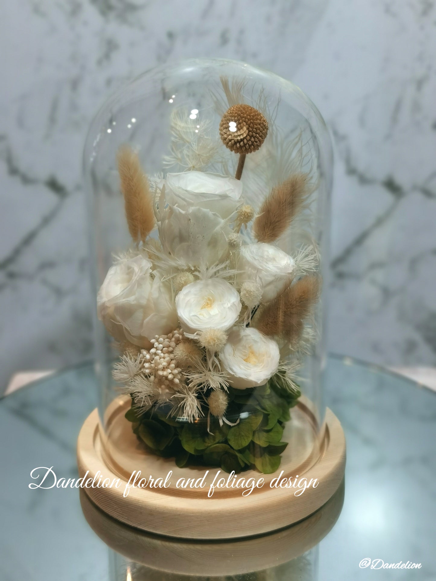 A preserved flower dome cloche arrangement with white flowers and dry plant elements, placed on a wooden stand.