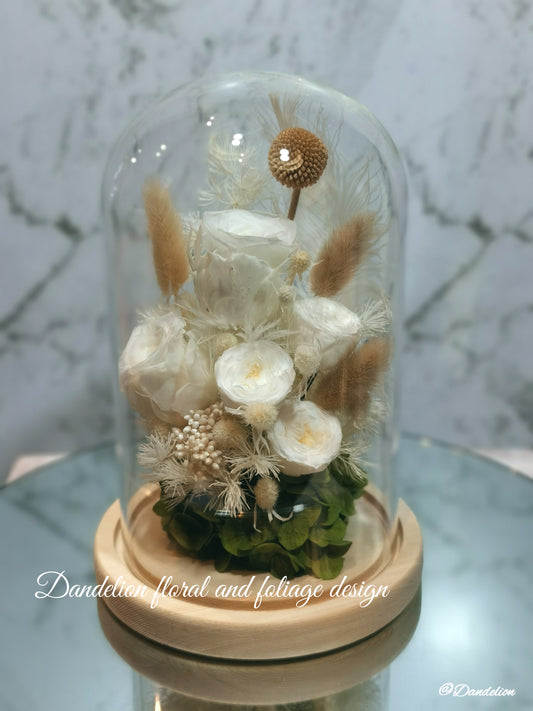 A preserved flower dome cloche arrangement with white flowers and dry plant elements, placed on a wooden stand.
