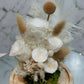 A preserved flower dome cloche arrangement with white flowers and dry plant elements, placed on a wooden stand.