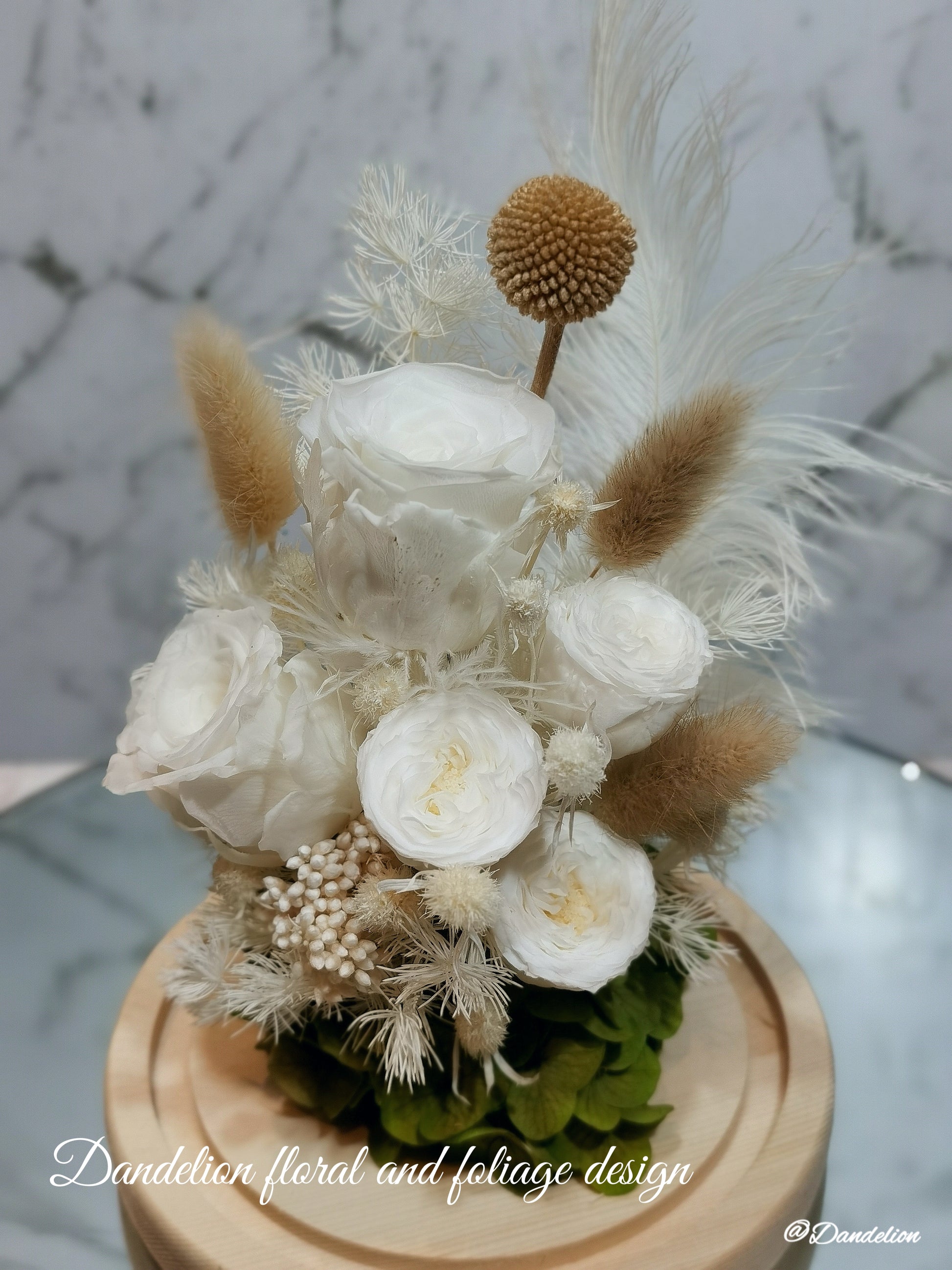 A preserved flower dome cloche arrangement with white flowers and dry plant elements, placed on a wooden stand.
