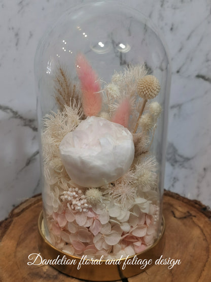 A handmade everlasting preserved white and pink flower dome cloche arrangement under a floral archway, displayed on a wooden stand. The dome is approximately 19cm in height and 10cm in diameter.