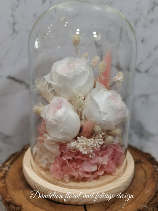 A handmade everlasting preserved white rose and pink flower dome cloche arrangement under a floral archway, displayed on a wooden stand.