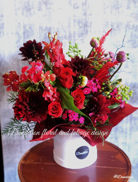 A flower box arrangement featuring a mix of red and pink flowers including roses and other botanicals, presented in a white container with a decorative red ribbon.