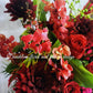 A flower box arrangement featuring a mix of red and pink flowers including roses and other botanicals, presented in a white container with a decorative red ribbon.