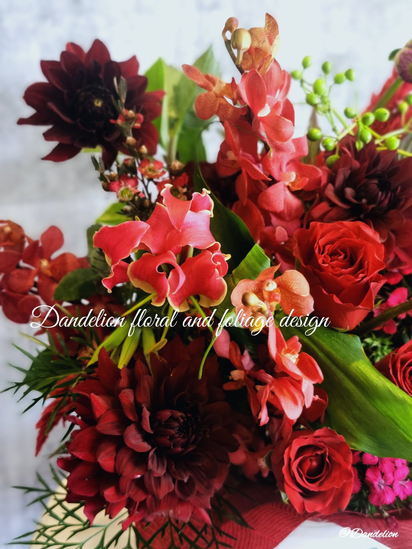 A flower box arrangement featuring a mix of red and pink flowers including roses and other botanicals, presented in a white container with a decorative red ribbon.