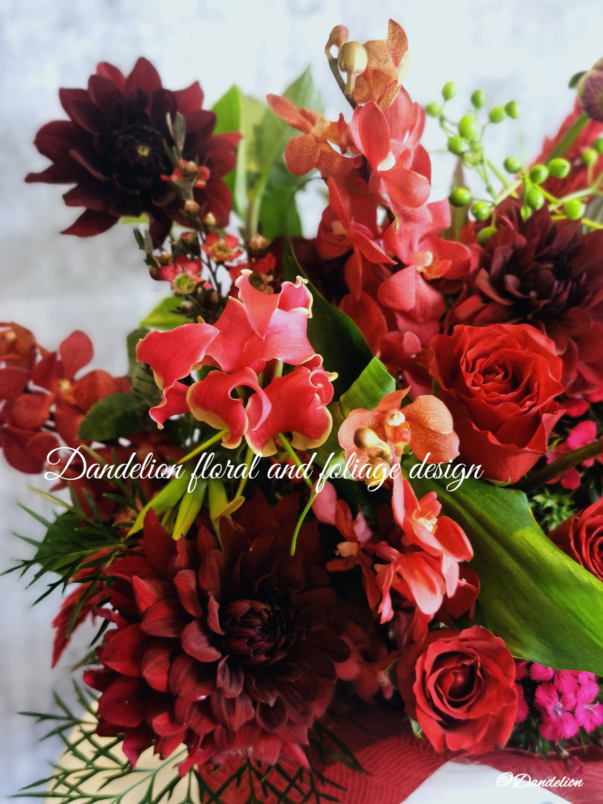 A flower box arrangement featuring a mix of red and pink flowers including roses and other botanicals, presented in a white container with a decorative red ribbon.