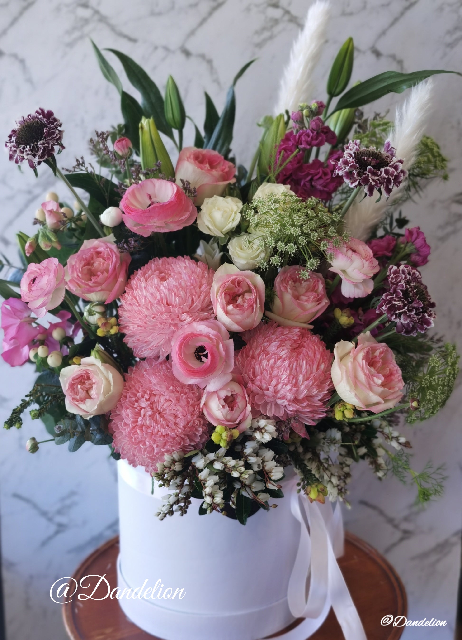 A round white hat box with a fresh flower arrangement consisting of pink, red and white flowers with green leaves.