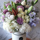 A round premium flat hat box arranged with a variety of flowers in shades of purple and white.