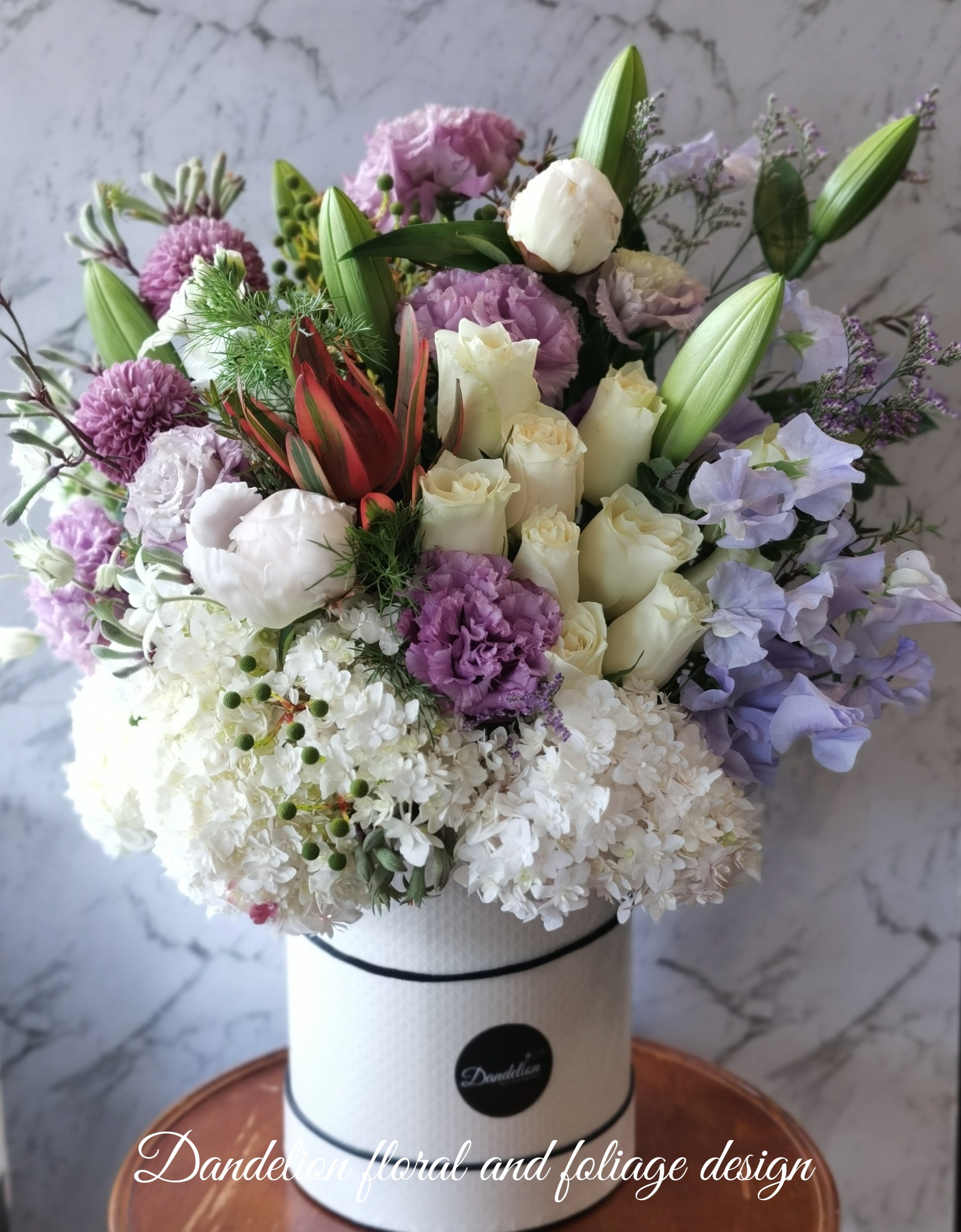 A round premium flat hat box arranged with a variety of flowers in shades of purple and white.