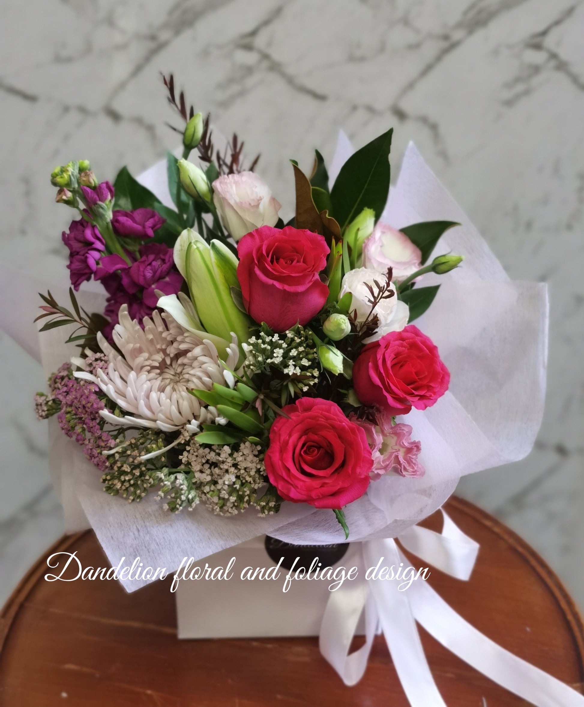 A floral arrangement featuring red roses, purple flowers, and green foliage, tied with white ribbons and placed in a white box.