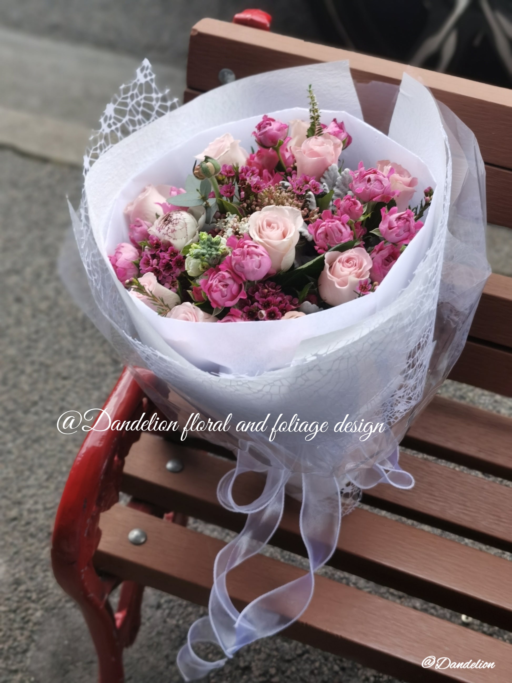 A mixed flower bouquet with white, pink roses, and soft mint green colors, wrapped and placed on a wooden bench.