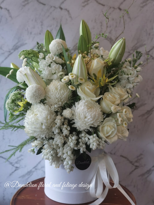 A round tall white hat box filled with white and green flowers, arranged white classic yet in a modern shape. Wedding, engagement or a sad occasion, this arrangement will fulfill what you are in need for.