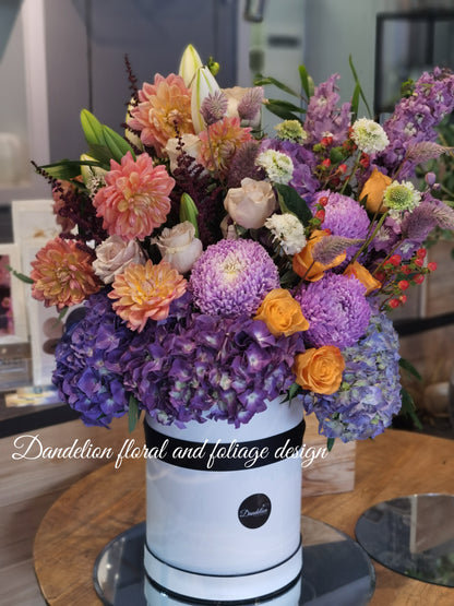 A bright colourful tall hat box arrangement using a mixture of roses, hydrangea, dahlia complimented with other seasonal flowers.