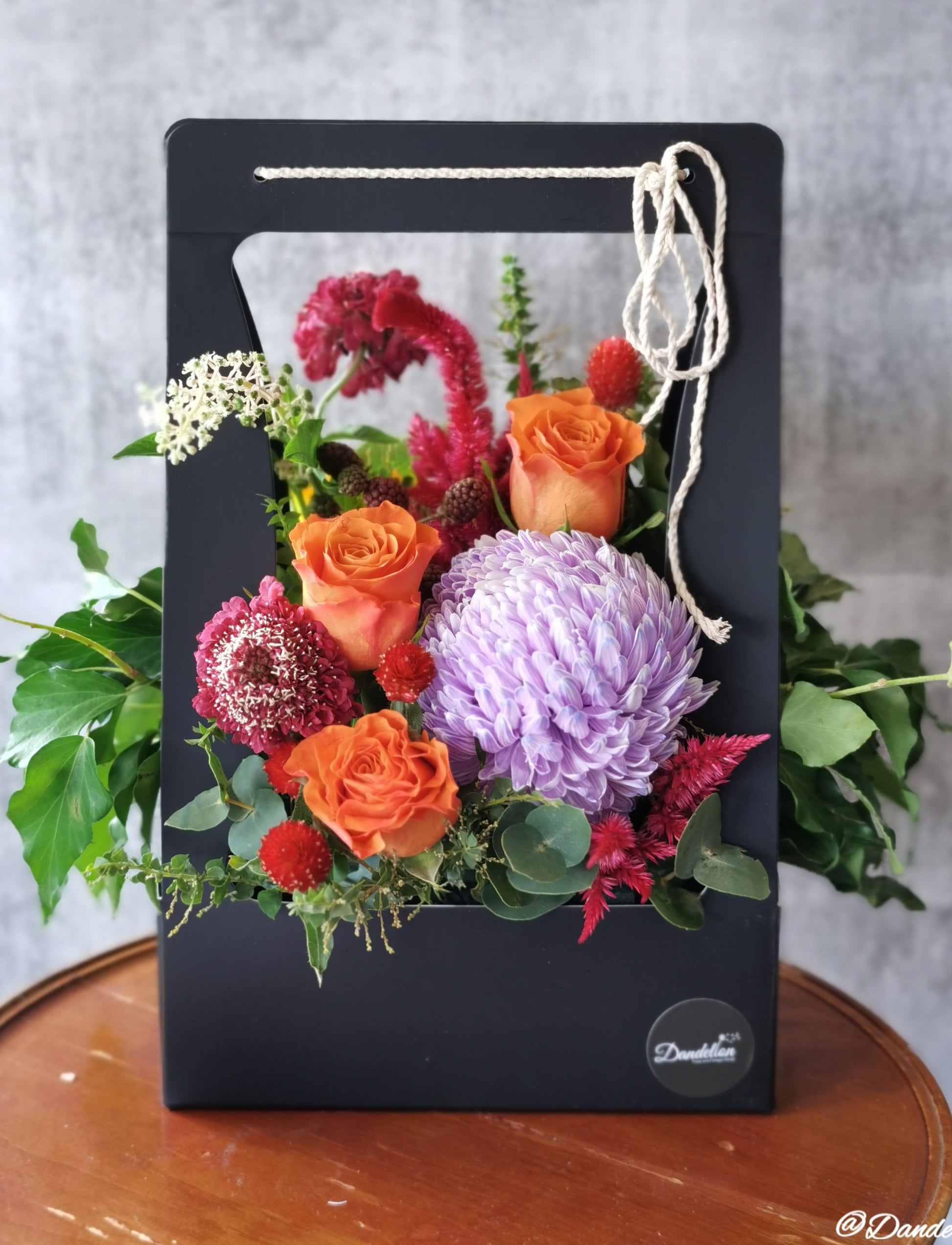 A floral carry box arrangement with a variety of flowers including a red carnation, orange roses, and green foliage, presented in a rectangular black box.