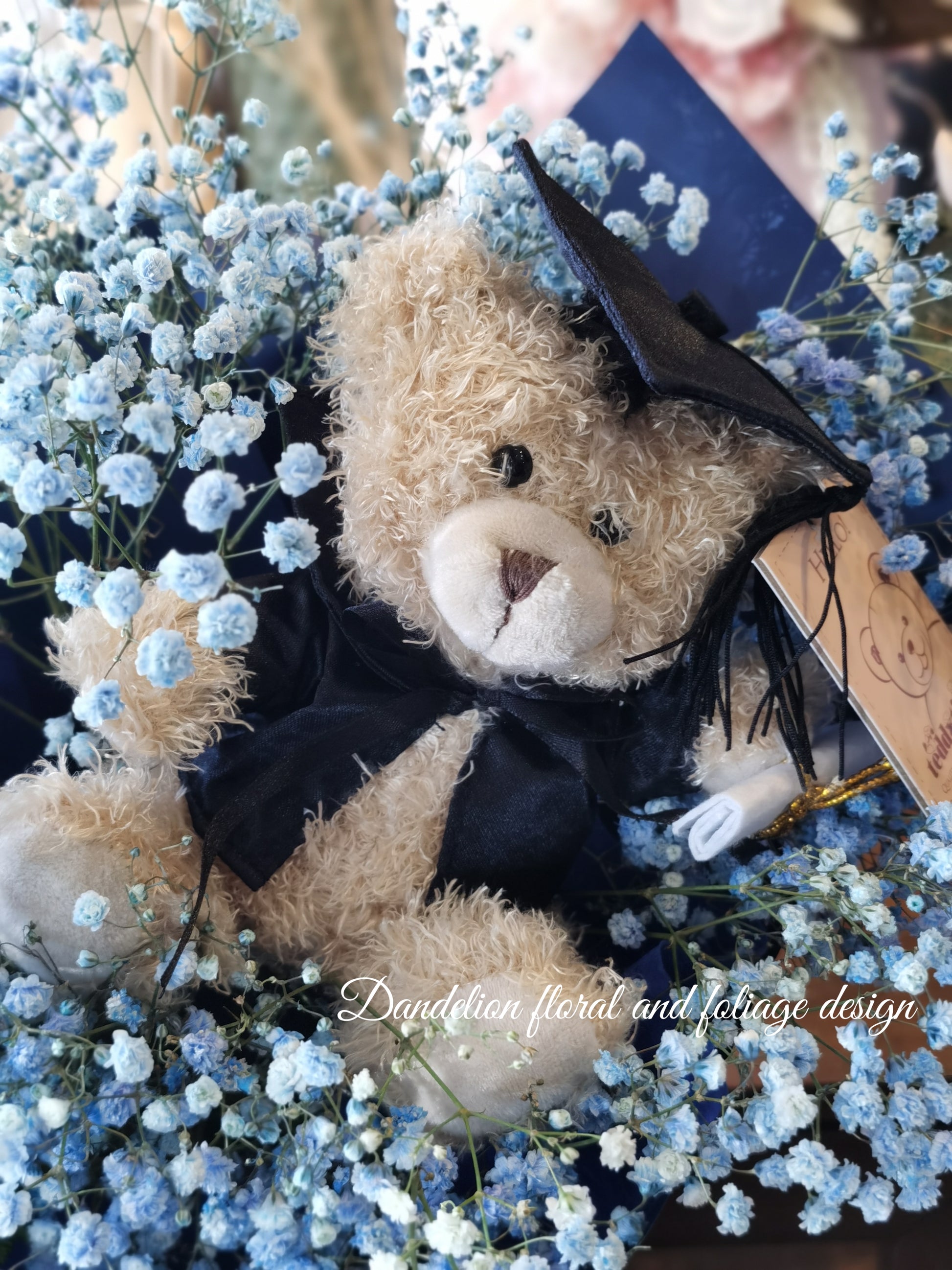 A bouquet of blue flowers with a teddy bear wearing a graduation cap and gown, surrounded by dark paper and a blue bow. 
Celebrate the achievement of your loved ones with beautiful graduation flowers. Find the perfect bouquet or arrangement to congratulate the graduate on their special day.