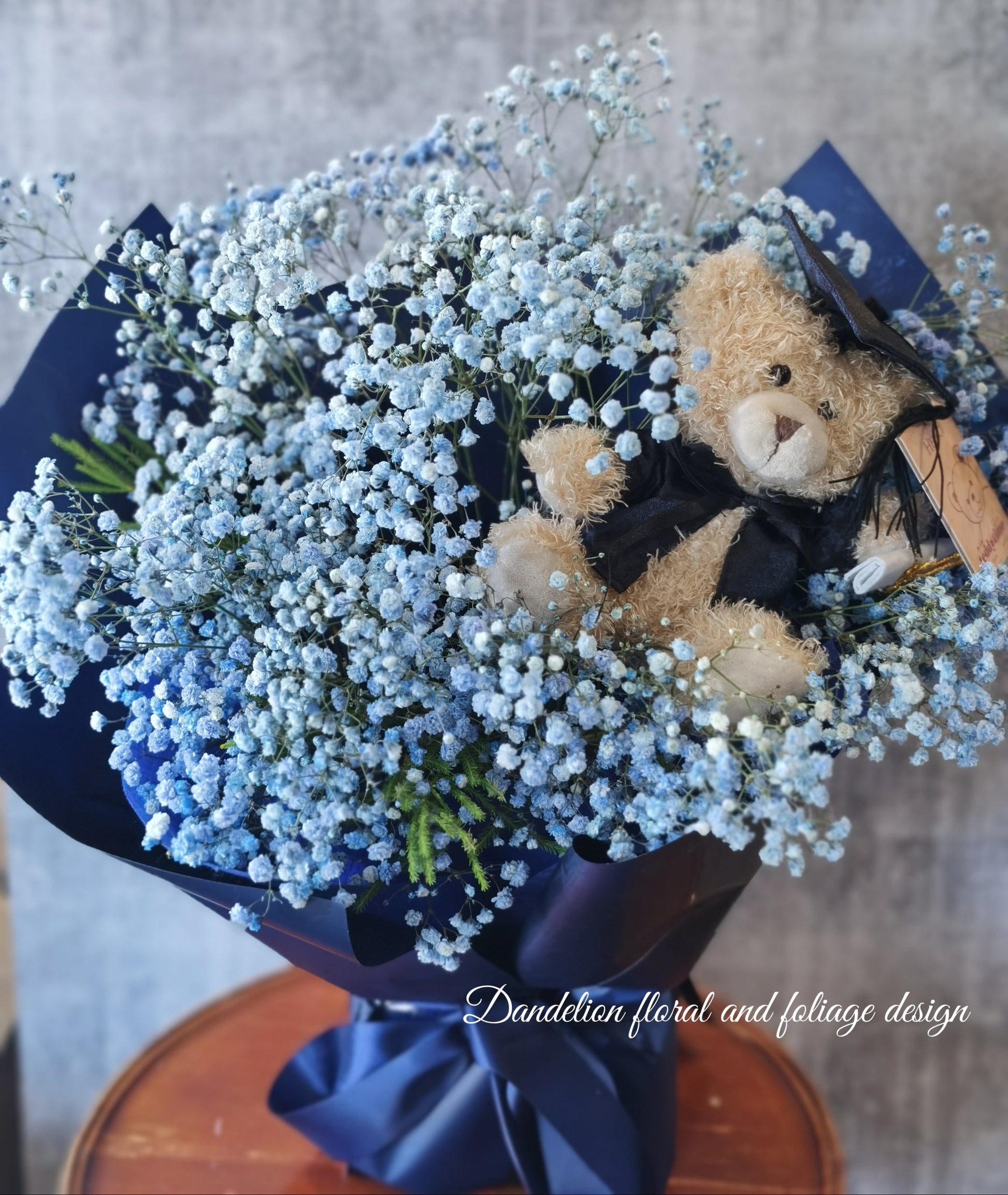 A bouquet of blue flowers with a teddy bear wearing a graduation cap and gown, surrounded by dark paper and a blue bow. 
Celebrate the achievement of your loved ones with beautiful graduation flowers. Find the perfect bouquet or arrangement to congratulate the graduate on their special day.
