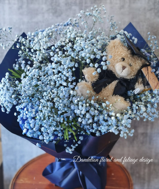 A bouquet of blue flowers with a teddy bear wearing a graduation cap and gown, surrounded by dark paper and a blue bow. 
Celebrate the achievement of your loved ones with beautiful graduation flowers. Find the perfect bouquet or arrangement to congratulate the graduate on their special day.