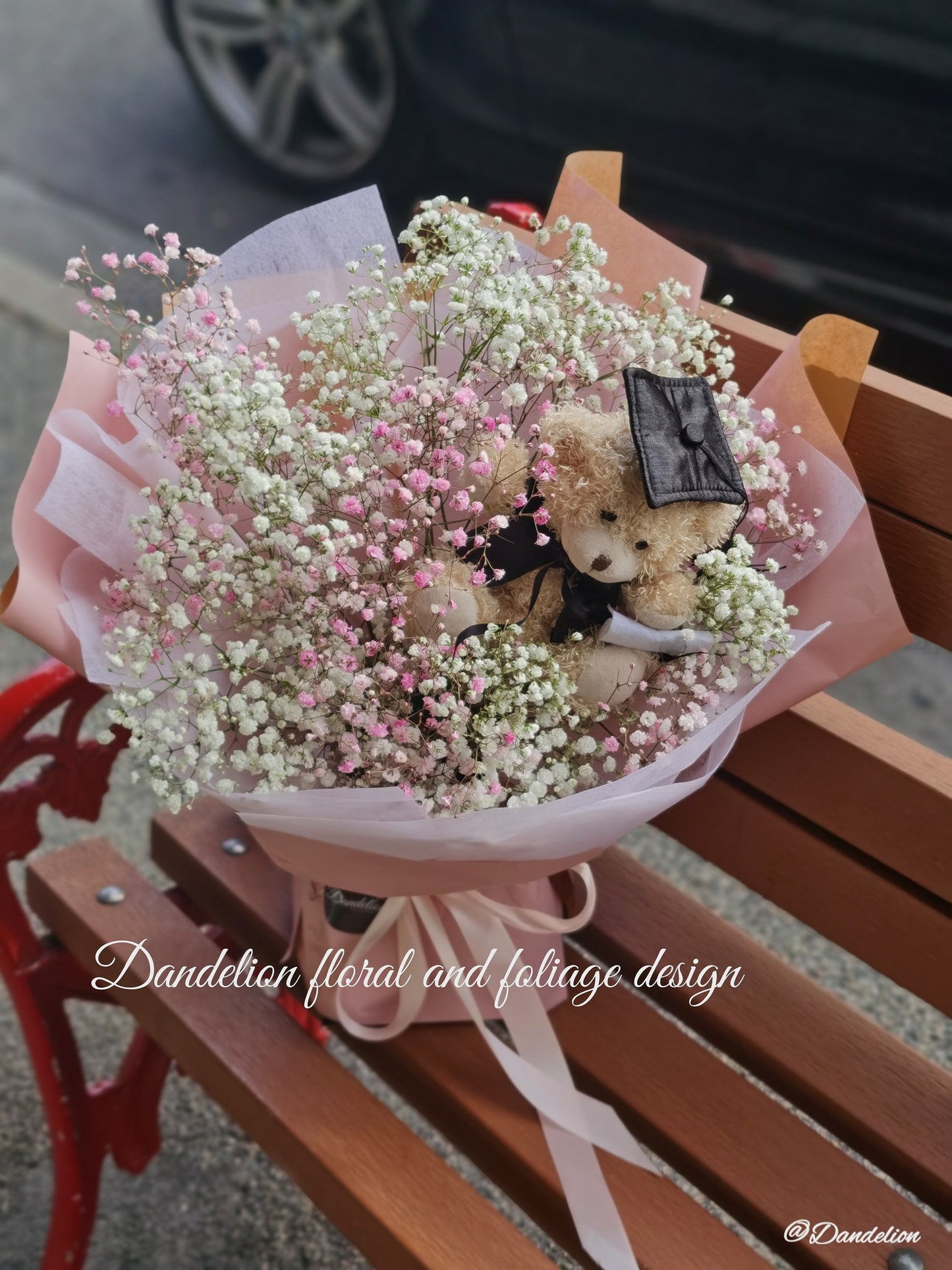 A bouquet of pink and white flowers with a teddy bear wearing a graduation cap and gown, surrounded by dark paper and a blue bow. 
Celebrate the achievement of your loved ones with beautiful graduation flowers. Find the perfect bouquet or arrangement to congratulate the graduate on their special day.