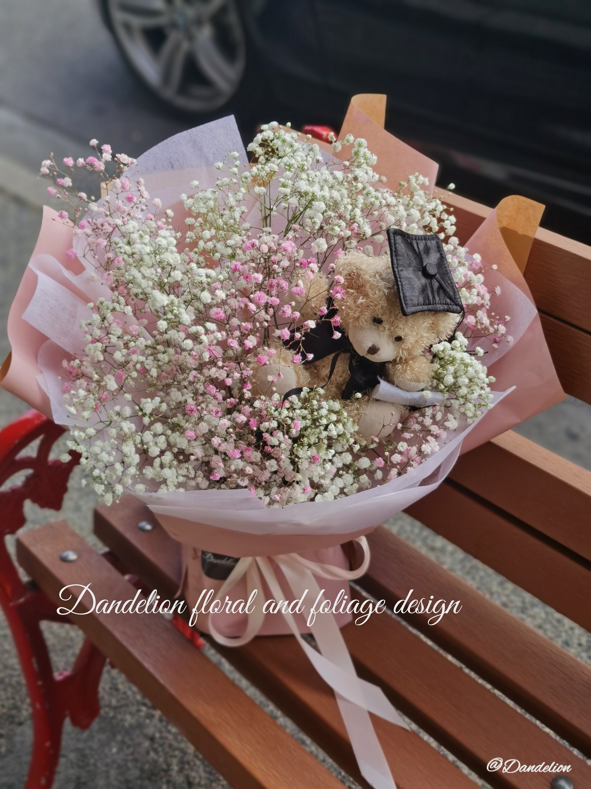 A bouquet of pink and white flowers with a teddy bear wearing a graduation cap and gown, surrounded by dark paper and a blue bow. 
Celebrate the achievement of your loved ones with beautiful graduation flowers. Find the perfect bouquet or arrangement to congratulate the graduate on their special day.