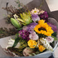 A bouquet of flowers with a mix of seasonal blooms and foliage, arranged and held by a mannequin. Bright colour bouquet using seasonal blooms complimented with seasonal foliage. Vibrant bouquet with sunflower, roses, lilies, natives and seasonal blooms in bright yellow and purple tones.