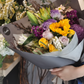 A bouquet of flowers with a mix of seasonal blooms and foliage, arranged and held by a mannequin. Bright colour bouquet using seasonal blooms complimented with seasonal foliage. Vibrant bouquet with sunflower, roses and seasonal blooms in bright yellow and purple tones.