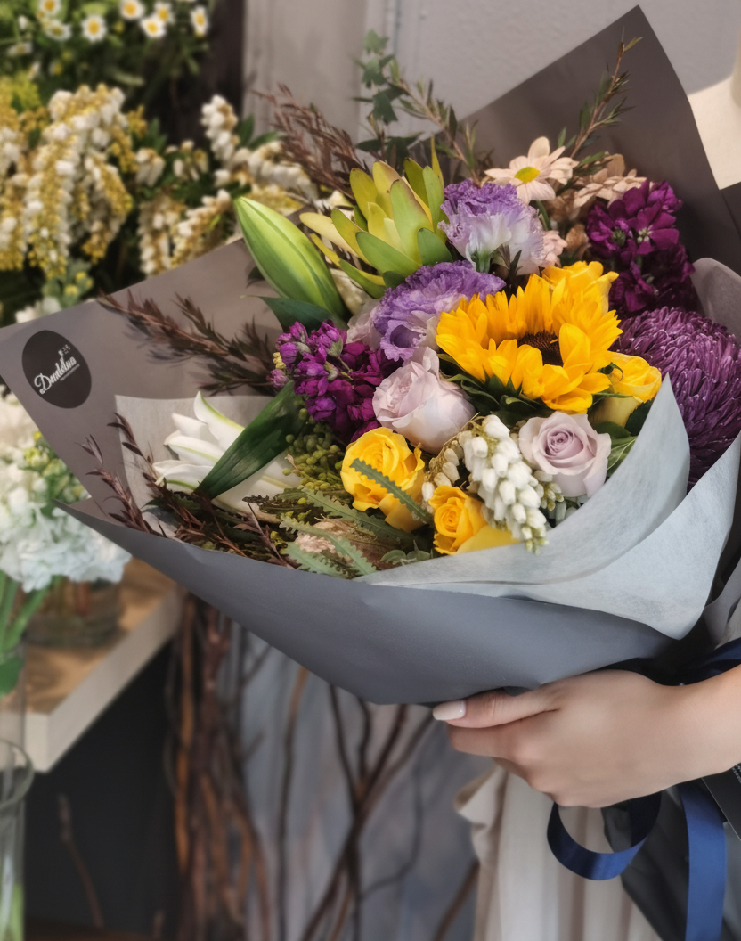 A bouquet of flowers with a mix of seasonal blooms and foliage, arranged and held by a mannequin. Bright colour bouquet using seasonal blooms complimented with seasonal foliage. Vibrant bouquet with sunflower, roses and seasonal blooms in bright yellow and purple tones.