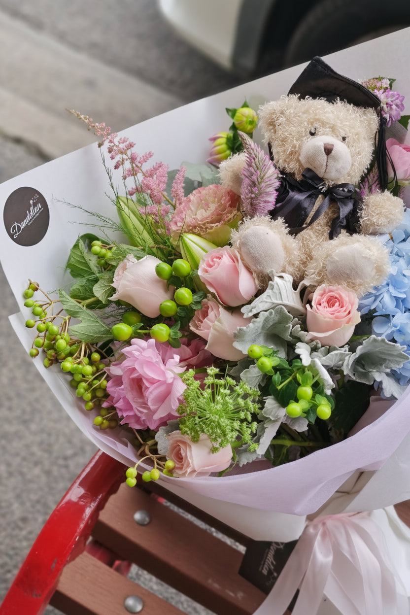 A graduation bouquet featuring a mix of pastel colored flowers and greenery, wrapped with a ribbon, alongside a plush toy bear. Lush bouquet using seasonal blooms in pastel tones complimented with seasonal greenery.
Our Graduation Flowers are the perfect way to send your best wishes and honour their hard work and dedication. With an added special touch our graduation bear - the perfect keepsake.